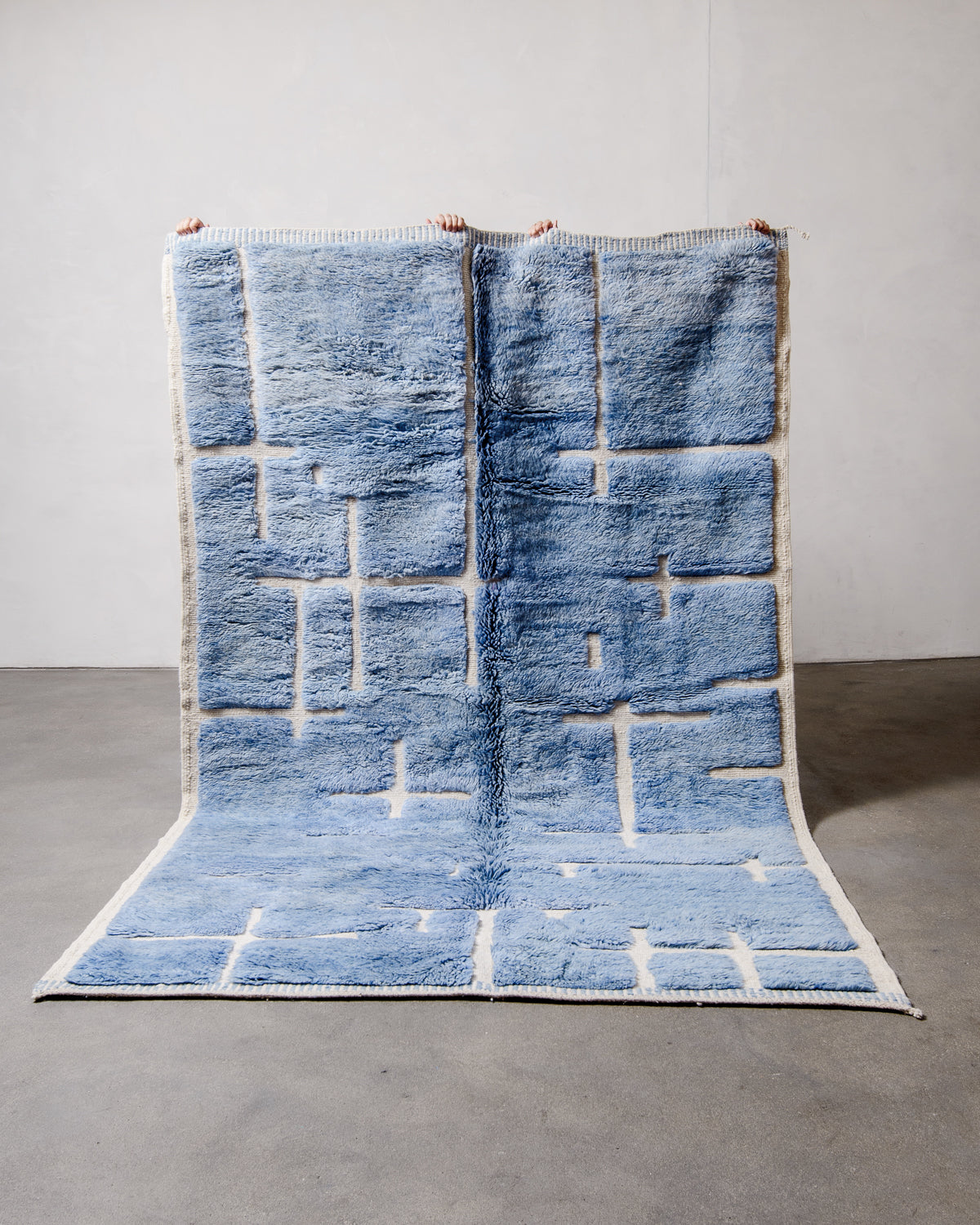 Berberlin Beni M'rirt Rug made of heavy wool pile in speckled denim blue and cream being held up in front of a white wall and concrete floor.